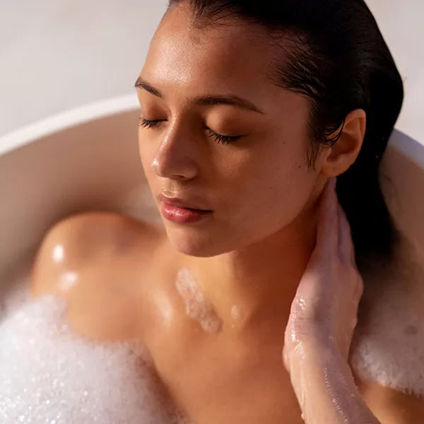 Como parte da experiência do Day Spa  amigas sintonia a foto mostra uma mulher dentro de uma banheira com espuma, s cabelos molhados e os olhos fechados. Ela repousa uma das mãos em seu pescoço