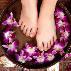 Como parte da experiência do Day Spa amigas a foto mostra pés mergulhados na água onde flutuam flores roxas em uma bacia de cor escura. Em volta da bacia há algumas conchas e pétalas de lor roxas