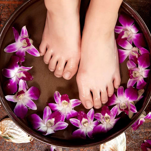 Como parte da experiência do Day Spa amigas a foto mostra pés mergulhados na água onde flutuam flores roxas em uma bacia de cor escura. Em volta da bacia há algumas conchas e pétalas de lor roxas