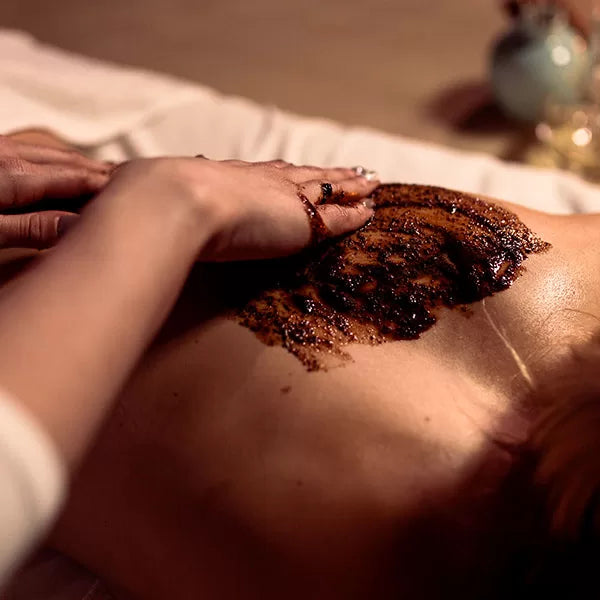 Capa do produto experiência esfoliação lama no speciale spa representado por uma foto que mostra as costas de uma mulher deitada de bruços. Uma mão, apoiada na altura da mulher, espalha uma pasta marrom granulada nas costas dela