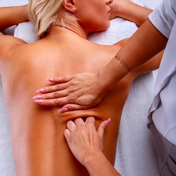 Como parte da experiência do Day Spa mãe e filha a foto mostra as costas de uma mulher deitada de bruços em cima de uma toalha branca e com a lateral do rosto apoiada nas mãos. Um par de mãos apertas a pele das costas dessa mulher