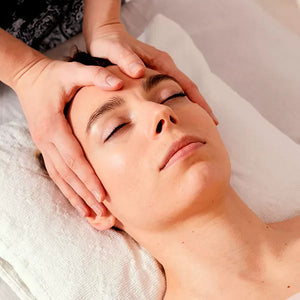 Como parte da experiência massagaem facial a foto mostra o rosto de uma mulher que está deitada em cima de uma toalha branca. Duas mãos seguram a cabeça dea, com os polegares em cima do meio da testa dela