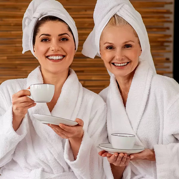 Como parte da experiência do Day Spa amigas a foto mostra duas mulheres vestindo roupões brancos e com os cabelos enrolados em toalhas brancas. As duas olham para a câmera e sorriem. Uma delas segura um pires branco com uma mão e na outra, um pires branco. A outra tem uma mão em cima da outra com as palmas para cima segurando um pires branco que apoia uma xícara branca
