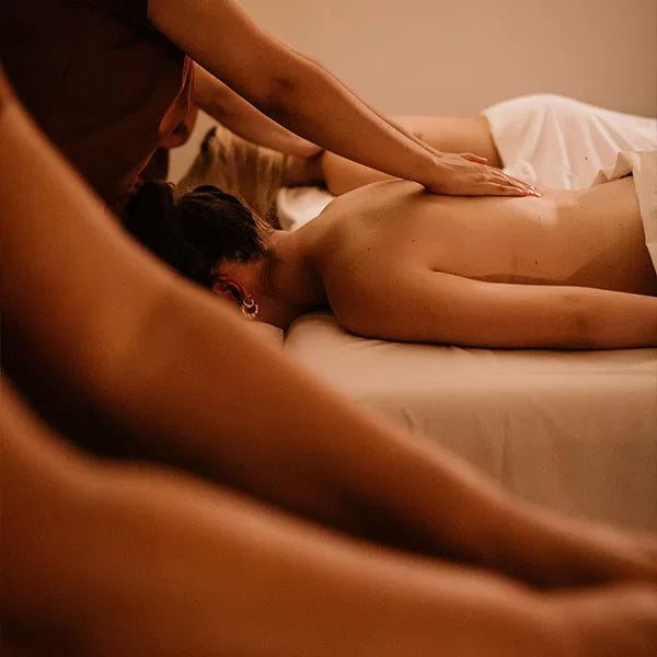 Como parte do produto day spa celebrate a foto mostra, em segunda plano, duas mulheres deitadas de bruços em macas e com toalhas brancas cobrindo seus quadris. Cada uma está recebendo massagem nas costas. Em primeiro plano, um par de braços, aparentemente também fazendo massagem, aparece