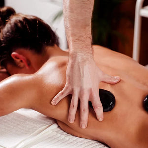 Como parte da experiência hot stones a foto mostra uma foto de uma mulher deitada de bruços sobre uma toalha branca. Uma mo segura uma pedra preta na lateral das costas