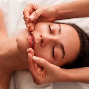Como parte da experiência massagaem facial a foto mostra o rosto de uma mulher que está deitada em cima de uma toalha branca. Duas mãos se apoiam nas bochechas, com a ponta dos dedos na lateral da boca