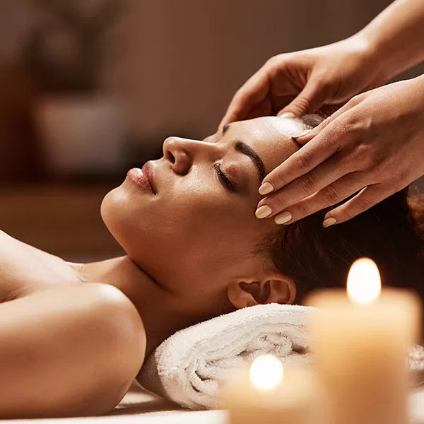 Como parte da experiência shiatsu facial a foto mostra a lateral o rosto de uma mulher deitada de olhos fechados sobre uma toalha branca. Duas mão seguram a lateral de sua cabeça