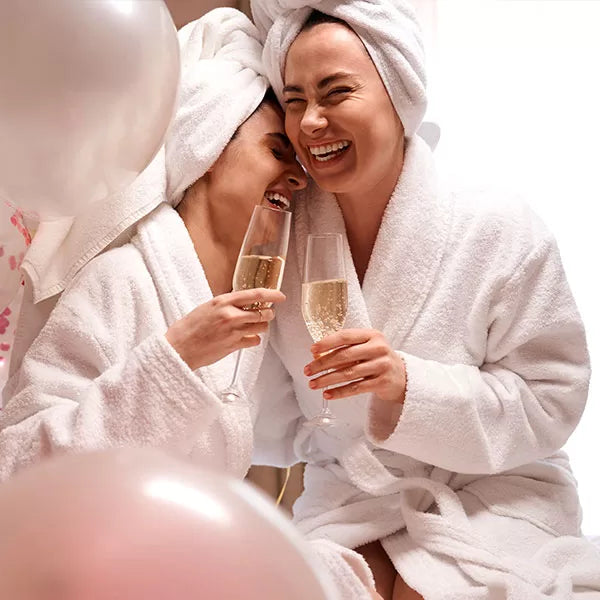 Como parte da experiência do Day Spa amigas sintonia a foto mostra duas mulheres vestindo roupões brancos, com toalhas brancas enroladas nos cabelos e segurando taças de champagne. Elas riem e tem os olhos fechados. Uma delas está com o rosto próximo ao pescoço da outra