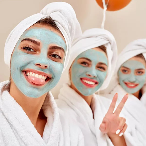 Como parte da experiência do Day Spa amigas a foto mostra três mulheres vestindo roupões brancos, com os cabelos enrolados em toalhas brancas e com um creme azul espalhado nos rostos. As três olham para a câmera e sorriem. Uma está com a ligua para fora e outra faz o sinal da paz com os dedos