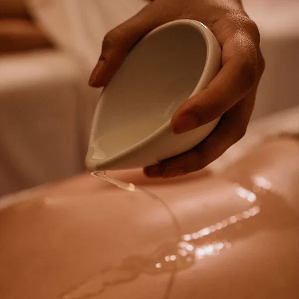 Como parte do produto day spa celebrate a foto mostra as costas de uma pessoa e, suspensa sobre ela, uma mão segura um recipinte branco de porcelaa, que está inclinado, derramando um líquido transparente nas  costas