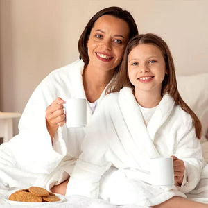 Como parte da experiência do Day Spa mãe e filha a foto mostra uma mulher e uma menina vestindo roupões brancos. Elas estã sentadas, olham para a câmera e cada uma segura uma xícara branca. A frente delas tem um prato branco com biscoitos