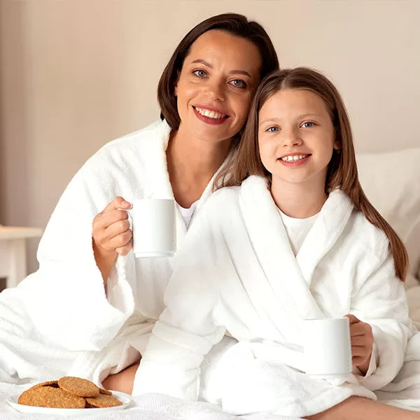 Como parte da experiência do Day Spa mãe e filha a foto mostra uma mulher e uma menina vestindo roupões brancos. Elas estã sentadas, olham para a câmera e cada uma segura uma xícara branca. A frente delas tem um prato branco com biscoitos