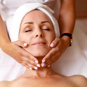 Como parte da experiência massagaem facial a foto mostra o rosto de uma mulher que está deitada em cima de uma toalha branca com os cabelas enrolados em uma toalha também branca. Duas mãos seguram a cabeça dela, com as mãos apoiadas na mandíbula