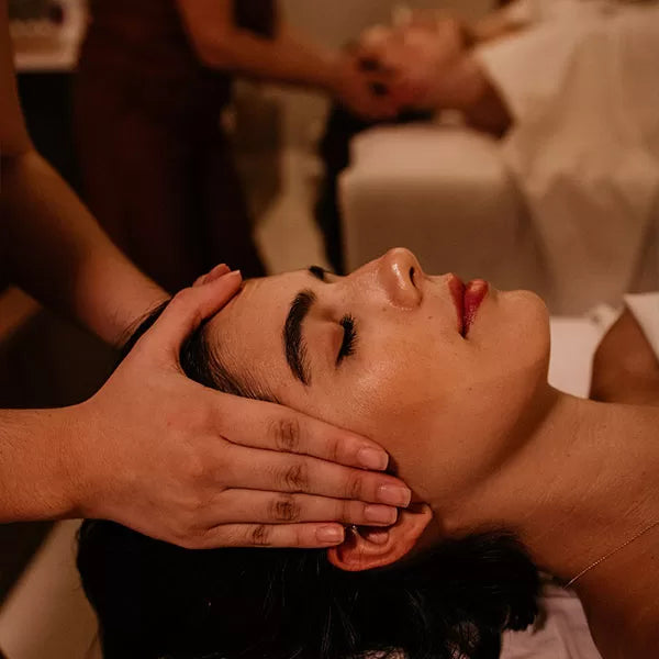 Como parte do produto day spa celebrate a foto mostra a lateral do rosto de uma mulher que está deitada e de olhos fechados. Duas mãos seguram as laterias de su cabeça. Ao fundo, desfocada, uma mulher também deitada, estpa com um lençol branco cobrindo seu corpo e duas mãos  também seguram as laterais de sua cabeça