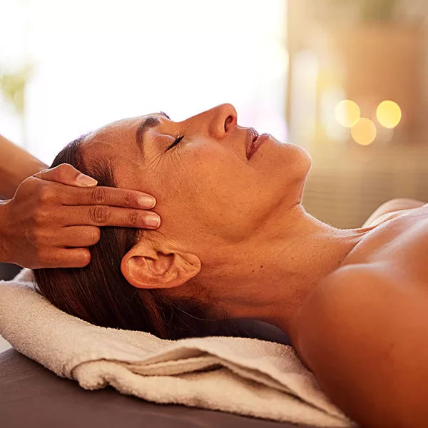 Como parte da experiência do Day Spa essence a foto mostra a lateral do rosto de uma mulher deitada que está de olhos fechados e a cabeça apoiada em uma toalha branca. Uma mão pressiona com a ponta de dois dedos suas têmporas