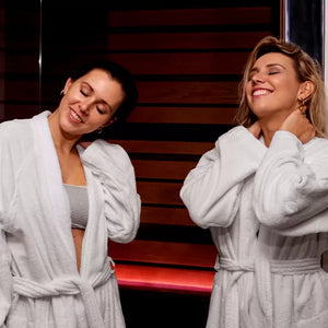 Como parte da experiência do Day Spa amigas a foto mostra duas mulheres vestidno roupões brancos, com olhos fechado e sorrindo. Uma delas segura a nuca com as duas mãos e a outra apoia uma das mão em seu próprio ombro