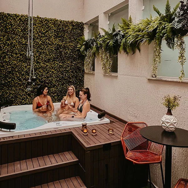Como parte do produto day spa celebrate a foto mostra três amigas dentro de uma jacuzzi, cada uma segurando uma taça de champagne e sorrindo. a jacuzzi é rodeada por uma estrutura de madeita e, apoiadas ao lado, há velas acesas. Na frente da estrutura de madeira, há uma mesa presa, com um vaso branco e uma planta em cima, e duas cadeiras vermelhas