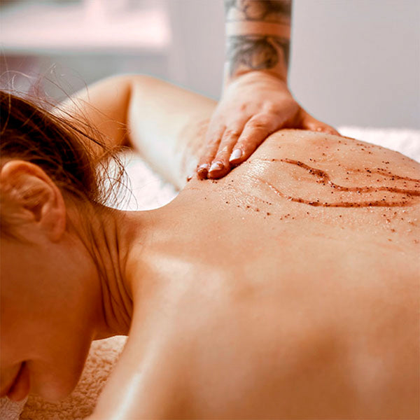 Como parte da experiência do Day Spa erva doce a foto mostra as costas de uma mulher deitada de bruços. Uma mão, apoiada na altura da mulher, espalha uma pasta marrom granulada nas costas dela