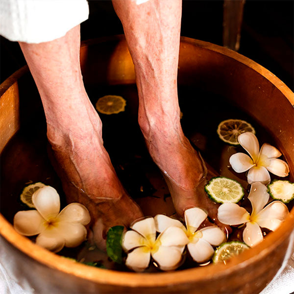 Como parte da experiência do Day Spa erva doce a foto mostra os pés de uma pessoas idosa submersos na água dentro de uma bacia de uma bacia. Na água flutuam flores brancas e rodelas de limão