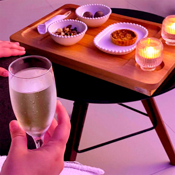 Como parte da experiência do Day Spa erva doce a foto mostra uma msa com tampo preto e pernas de madeira. Apoiada em cima dela está uma bandeja de madeira com talheres enrolados em um guardanapo branco, duas tigelas brancas com nozes e grão em uma e duas bolinhas marrom na outra. Em um prato oval branco há uma tortinha e dois recipientes de vidro com  duas velas acesas. Em primeiro plano, uma está apoiada no braço de uma poltrona e a outra segura uma taça de champagne