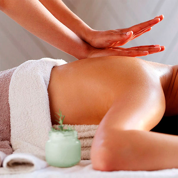 Como parte da experiência do Day Spa alecrim a foto mostra a lateral direita das costas de uma mulher que está deitada de bruços. Uma toalha marrom e branca está cobrindo metade de suas costas e seu peito está apoiado em uma toalha bege. Ao lado de seu corpo há um frasco de vidro com um creme branco e um ramo de alecrim espetado no creme. Uma mão direita está tocando as costas da mulher enqanto uma mão esquerda está apoiada em cima da direita, com os dedos levemente inclinados para cima