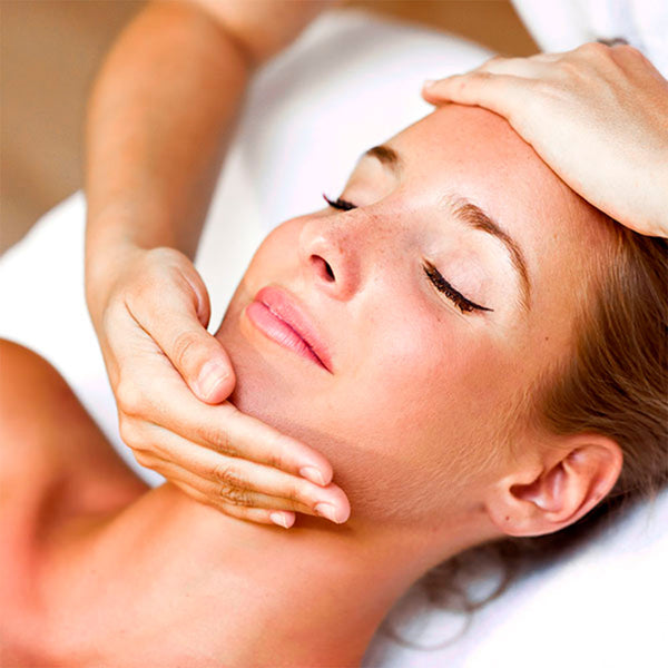 Como parte da experiência do Day Spa alecrim a foto mostra o rosto de uma mulher deitada em cima de uma toalha branca. Seus olhos estão fechados e sua expressão está serena. Uma mão direita está cobrindo o queixo da mulher e a esquerda está apoiada em sua testa