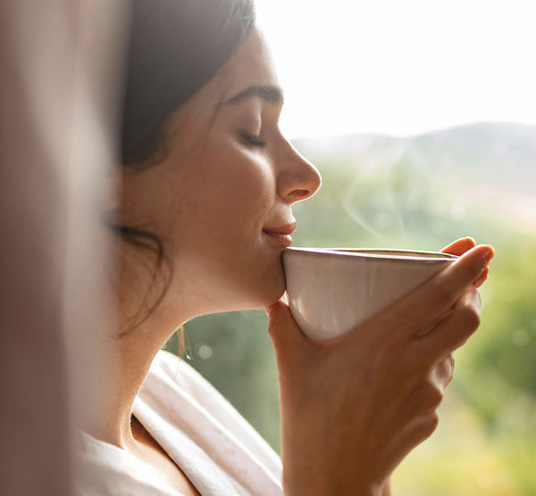 Como parte da experiência do Day Spa balance a foto mostra uma mulher de frente para uma janela, segurando com as duas mãos uma xícara branca e a encostando no queixo