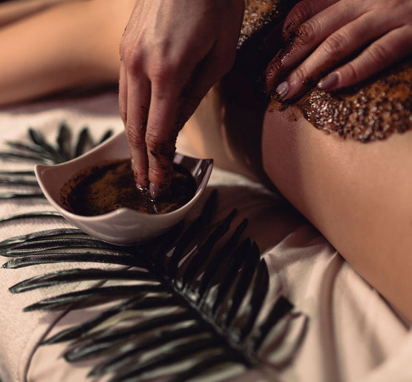 Como parte da experiência do Day Spa balance a foto mostra duas mãos e parcialmente as costas de uma mulher. Uma das mãos está tocando com as pontas dos dedos uma mistura marrom granulada que está dentro de um pote branco pousado ao lado das costas e em cima de uma folha verde. a outra mão repousa em cima das costas, onde já existe essa mistura espalhada