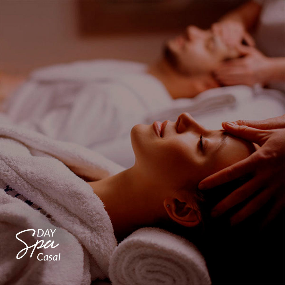 Capa do produto day spa casal no speciale spa representado porum homem e uma mulher, cada um deitado em uma maca, vestindo roupões brancos e recebendo massagem na cabeça. O homem está ao fundo da foto, desfocado, enquanto a mulher está em primeiro plano
