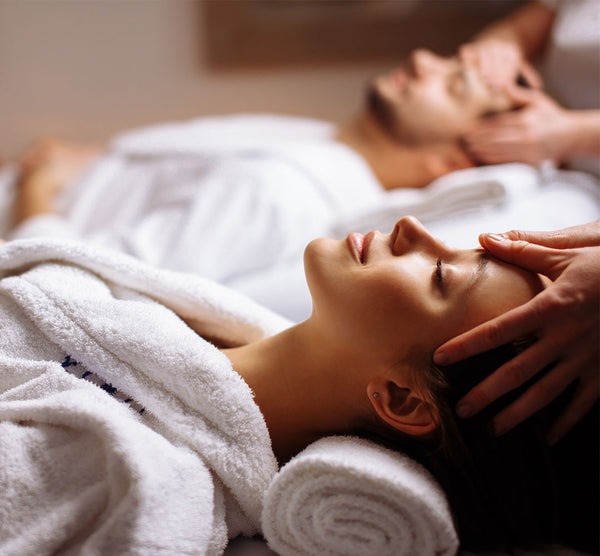 Como parte da experiência do Day Spa casal harmonia a foto mostra um homem e uma mulher, cada um deitado em uma maca, vestindo roupões brancos e recebendo massagem na cabeça. O homem está ao fundo da foto, desfocado, enquanto a mulher está em primeiro plano