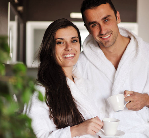 Como parte da experiência do Day Spa casal harmonia a foto mostra um um homem e uma mulher, abraçados de lado um ao outro, olhando para câmera e sorrindo. Cada um segura uma xícara branca e vestem roupões brancos