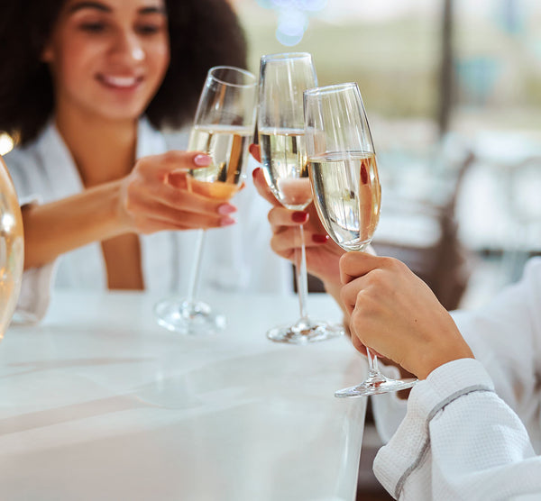 Como parte da experiência do Day Spa Encontro para amigas a foto mostra um brinde  com três taças de champagne  sendo seguradas por três mãos femininas. Uma das mulheres aparece desfocada ao fundo 