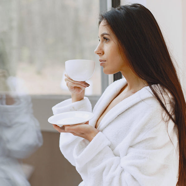 Como parte da experiência do Day Spa lavanda a foto mostra uma mulher morena, vestindo um roupão branco, olhando por uma janela, segurando em uma das mãos um pires branco e na outra uma xícara branca, que ela segura na altura de sua boca