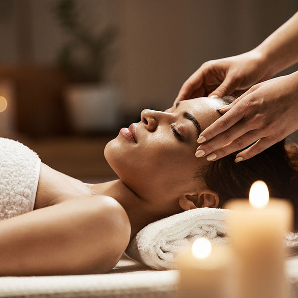 Como parte da experiência do Day Spa lavanda a foto mostra a lateral esquerda uma mulher deitada, coberta até a altura do peito com uma toalha branca e com a cabeça apoiada a uma toalha também branca. Seus cabelos estão repousados para trás e as duas mãos estão apoiadas em suas têmporas