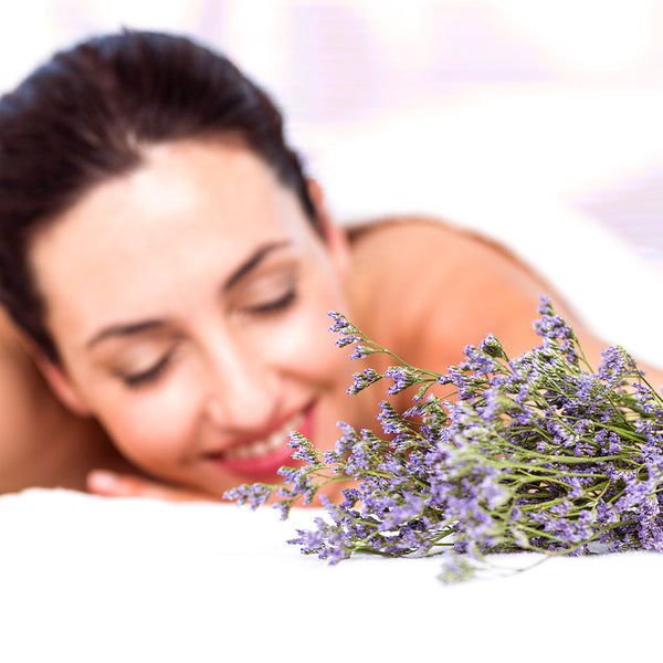 Como parte da experiência do Day Spa lavanda a foto mostra ramos de uma flor roxa pequena apoiada sobre uma toalha branca. Ao fundo, desfocada, uma mulher está deitada de bruços com o rosto voltado para a câmera e a cabeça ligeiramente inclinada para a direita e o queixo apoiado em cima de sua mão. Ela está com os olhos fechados e sorrindo