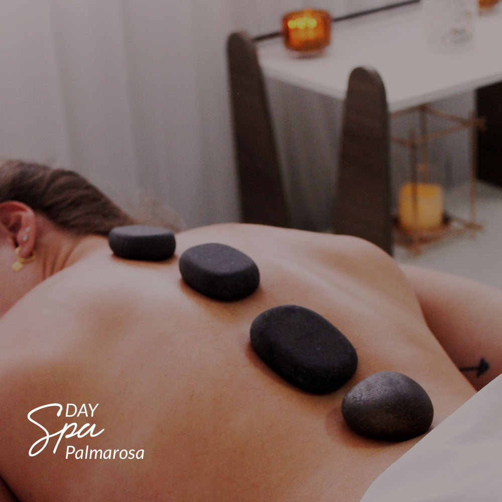 Capa do produto day spa palmarosa no speciale spa representado por uma foto de uma mulher deitada de costas. Sua cabeça está voltada para cima do quadro e seu rosto, que não aparece, está virado para a esquerda. Quatro pedras estão alinhadas na linha da coluna da mulher e, ao fundo, desfocado, há um aparador branco com pernas marrons que apoia uma vela acesa. Embaixo do aparador também há uma vela grande acesa