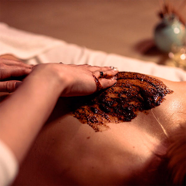 Como parte da experiência do Day Spa palmarosa a foto mostra as costas de uma mulher com uma pasta marrom granulada sendo espalhada por duas mãos