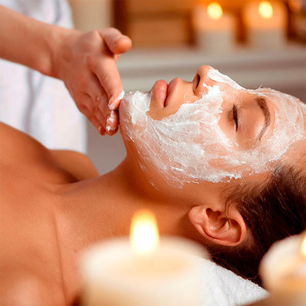 Como parte da experiência do Day Spa palmarosa a foto mostra a lateral esquerda do rosto de uma mulher deitada. Seu rosto está coberto por uma pasta branca que está sendo espalhada por uma mão. em primeiro plano, desfocada, há uma vela acesa