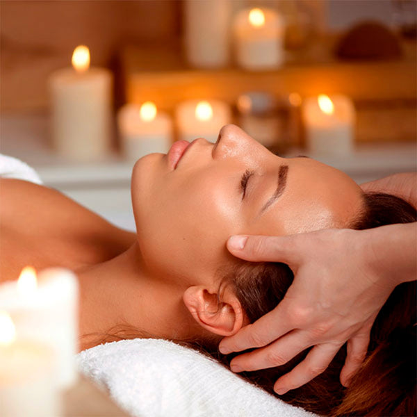Como parte da experiência do Day Spa palmarosa a foto mostra a lateral esquerda do rosto de uma mulher deitada. Uma mão esquerda está apoiada na lateral de sua cabeça e em primeiro plano, desfocada, há uma vela acesa, assim como ao fundo, também desfocadas, há diversas veas acesas