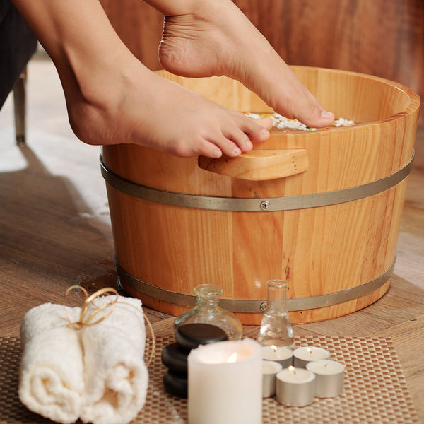 Como parte da experiência do Day Spa speciale a foto mostra pés apoiados na borda de uma bacia de madeira. A bacia está com água e umas pételas brancas. Ao lado da bacia, em primeiro plano, há uma toalha branca dobrada e amarrada com uma fota fina bege, cinco velas pequenas e uma grande acesas, dois frascos de vidro e três pedras redondas empilhadas