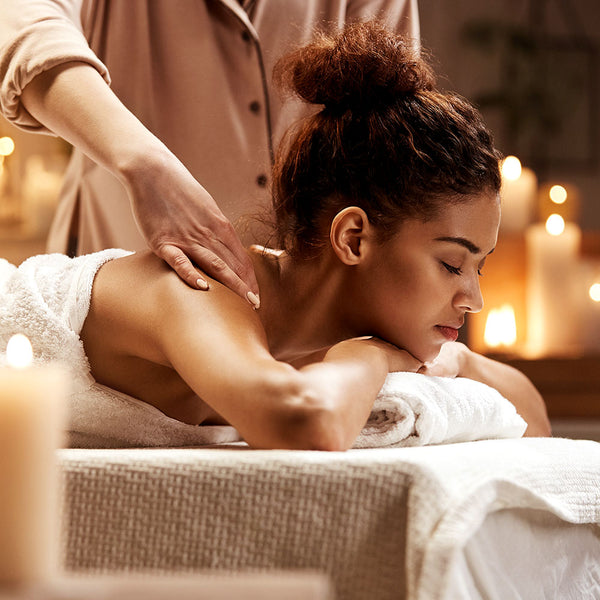 Como parte da experiência do Day Spa speciale a foto mostra a lteral direita do troco e cabeça de uma mulher enrolada em uma toalha branca, que está deitada de bruços em uma maca forrada também com uma toalha branca, com queixo apoiado na parte de trás de suas mãos e que por sua vez, estão apoiadas em uma toalha branca. Ela está com os olhos fechados e uma expressão serena. Atrás dela, há uma pessoa em pé, massageando suas costas. Ao redor delas, há diversas velas acesas