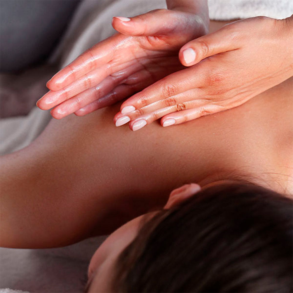 Capa do produto experiência massa no speciale spa representado pela foto mostra a lateral direita das costas de uma mulher que está deitada de bruços. Uma toalha marrom e branca está cobrindo metade de suas costas e seu peito está apoiado em uma toalha bege. Ao lado de seu corpo há um frasco de vidro com um creme branco e um ramo de alecrim espetado no creme. Uma mão direita está tocando as costas da mulher enqanto uma mão esquerda está apoiada em cima da direita, com os dedos levemente inclinados para cima
