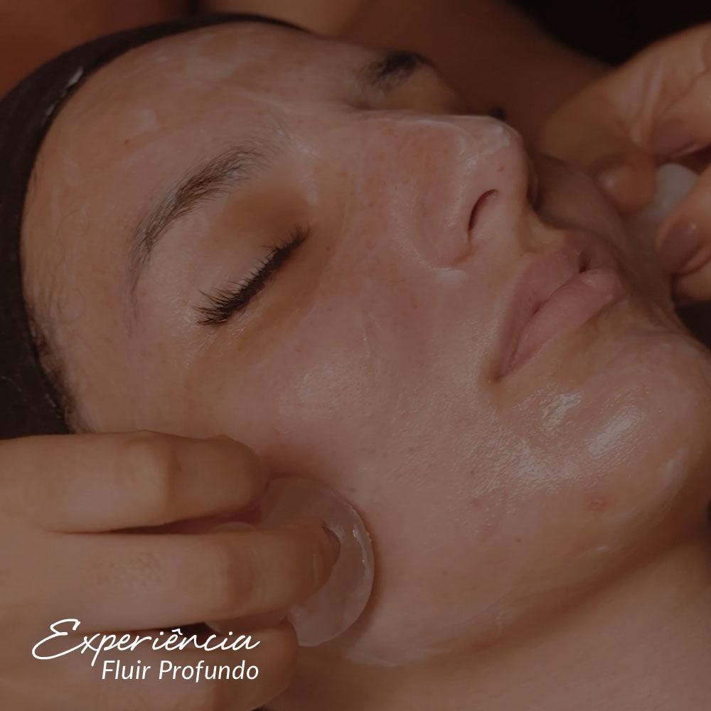 Capa do produto experiência fluir profundo com a imagem do rosto de uma mulher deitada de olhos fechados. ela está com uma faixa preta na linha do cabelo na testa e um creme branco está espalhado no rosto. duas mão estão passando objetos translúcidos rosados em cada um dos lados da face