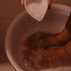 Dois pés estão submersos na água em uma bacia branca. Uma mão segurando um recipiente de cerâmica branco paira sobre a bacia e derruba sal grosso na água