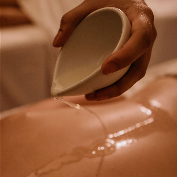 As costas nuas de uma pessoa deitada de bruços. Uma mão segurando um recipiente de cerâmica branco derrama um líquido transparente sobre as costas