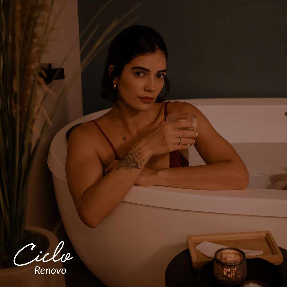 Capa do produto ciclo renovo. Uma mulher, que encara a câmera, segurnado um copo de vidro e de biquíni, submersa na água em uma banheira branca. A parte de cima de seu corpo está fora da água e seus braços quase se cruzam na bosrda da banheira. Ao lado, há uma pequena mesa com uma bandeja de madeira com um guardanapo e uma vela acesa. Atrás da banheira, há uma grande planta verde.