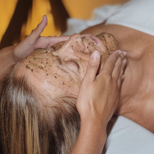 Imagem do rosto de uma mulher deitada de olhos fechados e com um creme amarronzado espalhado nele. Duas mãos estão posicionadas uma de cada lado do rosto