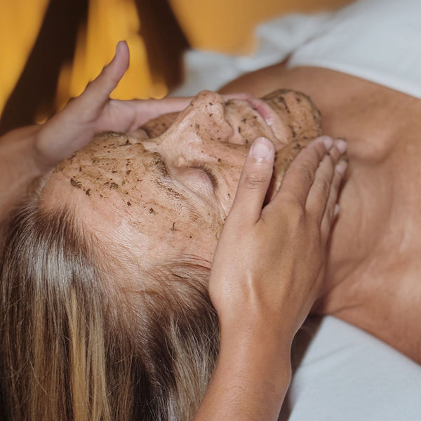 Imagem do rosto de uma mulher deitada de olhos fechados e com um creme amarronzado espalhado nele. Duas mãos estão posicionadas uma de cada lado do rosto