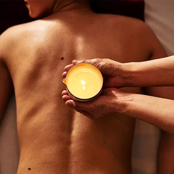 Capa do produto experiência candle massage no speciale spa representado por duas mão em cima das costas de uma pessoa deitada de bruços. Uma das mãos está apoiada nas costas  com a palma para cima, em concha. A outra mão segura um recipiente de vidro com uma vela acesa dentro e inclinada em cima da outra mão 
