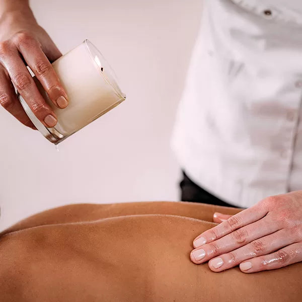 Como parte da experiência candle massage a foto mostra duas mão em cima das costas de uma pessoa deitada de bruços. Uma das mãos está apoiada nas costas  e a outra mão segura um recipiente de vidro com uma vela acesa dentro e inclinada em cima das costas